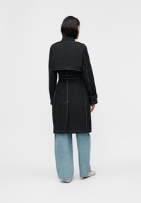 Γυναίκα που φοράει μαύρο παλτό με ζώνη και λευκές ραφές, ανοιχτό μπλε τζιν παντελόνι σε φαρδιά γραμμή και μπεζ παπούτσια, όρθια με την πλάτη προς τα εμπρός.