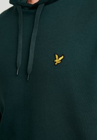 Sudadera de color verde oscuro con tela texturizada, que presenta un pequeño logo bordado de un pájaro amarillo en el lado izquierdo del pecho y detalles con cordones.