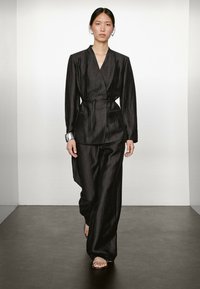 Schwarzer strukturierter Stoffanzug mit einem Wickel-Blazer und Gürtel sowie weit geschnittenen Hosen. Silberne Armree, minimalistisches Design.