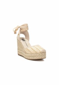 Sandalia de cuña con una parte superior de textil tejido en beige claro, puntera redonda y suela de yute trenzado. Presenta una correa de tela en el tobillo atada en un lazo.