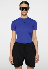 Blå, figursydd kortärmad skjorta med en svart logotyp på bröstet, tillsammans med svarta lösa shorts med elastisk midja och snörning.