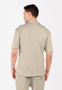 Man met een beige shirt met korte mouwen en een bijpassende broek, gezien vanaf de achterkant tegen een witte achtergrond.