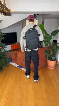 Black backpack with multiple external pockets, rectangular design, adjustable straps, and a smooth texture, carried by a person in a casual setting.