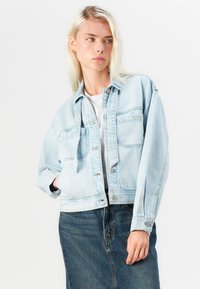 Light blue denim jacket with a cropped design, button-up front, two chest pockets, and cuffed sleeves, paired with a white shirt and dark skirt.