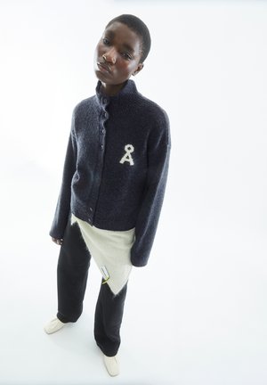 Marineblauwe gebreide cardigan met hoge kraag, knoopsluiting en witte letter "A" embleem. Gecombineerd met een beige wikkelpantalon en zwarte broeken.