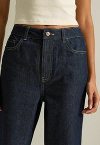 Donna che indossa jeans a vita alta blu scuro e un top corto bianco, mostrata dalla vita fino a metà coscia su uno sfondo neutro.