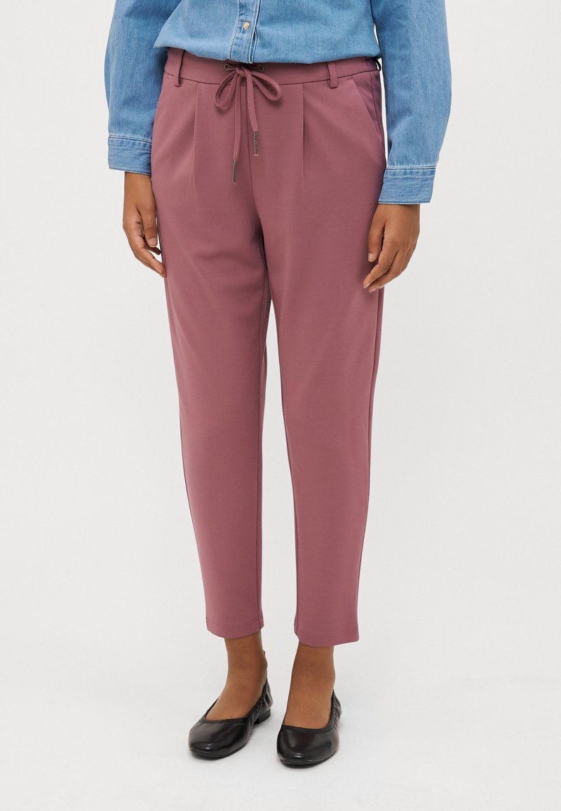 Femme portant un pantalon mauve à cordon, des ballerines noires et une chemise en jean bleu clair sur un fond blanc uni.