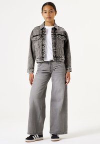 Grijze denimjas met voorzakken, hoge kraag en rafelige randen. Gepaard met grijze jeans met wijde pijpen en zwarte sneakers met witte strepen.