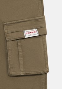 Khaki stoff med cargo-lomme med klaff og hvit etikett med teksten "VINGINO THE MAKING OF ICONS EST. 1997" sydd på lommens klaff.