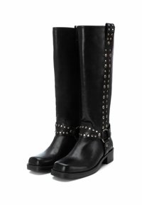 Botas de cuero negro hasta la rodilla con un eje recto, acentos de tachuelas y una correa con hebilla alrededor del tobillo. Suela plana, acabado texturizado.