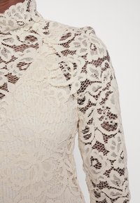 Cream lace top with a high collar and long sleeves, featuring floral patterns and a sheer texture, highlighting intricate detailing.