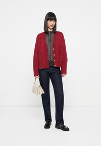 Rød kabelstrikket cardigan med gullknapper, brukt over en svart prikkete skjorte, kombinert med mørke denim jeans og svarte loafers, bærende på en beige håndveske.