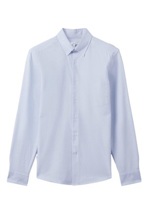 Nærbillede af en person iført en lyseblå skjorte med knapper, med knappet krave og brystlomme, mod en neutral baggrund.