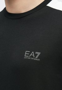 Fekete pamut póló kerek nyakkivágással. Bal oldalon a mellkason ezüst EA7 Emporio Armani logóval és fehér részletekkel a vállakon.