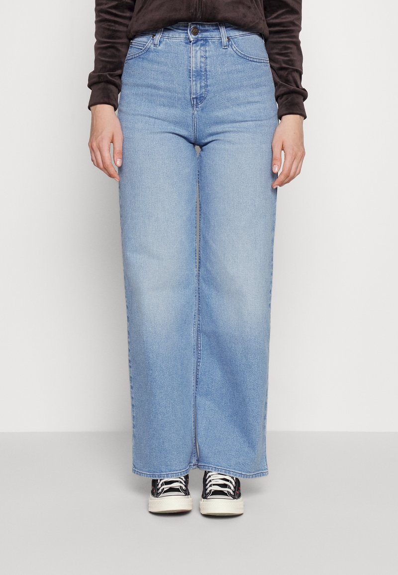 Personne portant un jean bleu clair à jambes larges, un haut noir à manches longues et des baskets noires et blanches, debout devant un fond uni.