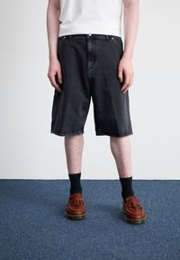 Black denim shorts with a relaxed fit, featuring a button fly and pockets. Paired with brown shoes and black socks on a blue carpet.