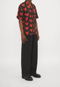 Uomo che indossa una camicia nera a maniche corte con bottoni e motivi a cuore di diavolo rossi, pantaloni neri larghi e stivali in pelle nera.