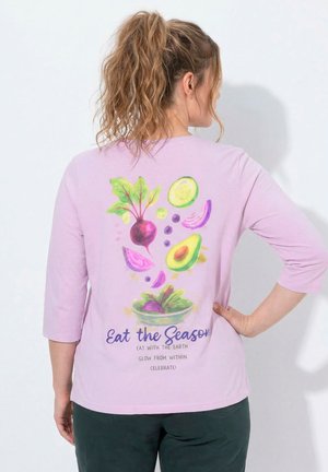 Frau mit Pferdeschwanz, die ein hellviolettes Hemd trägt, das auf der Rückseite ein buntes Gemüse-Design und den Text "Eat the Season" zeigt.
