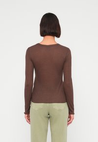 Καφέ μακρυμάνικη μπλούζα με στρογγυλή λαιμόκοψη, εφαρμοστή γραμμή και απαλή υφή, συνδυασμένη με ανοιχτό πράσινο παντελόνι που διαθέτει λεπτομέρειες σε πίσω τσέπη.