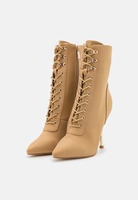 Bottes beige montant jusqu'au genou avec un bout pointu, un laçage à l'avant et des éléments métalliques dorés ; dotées d'un talon stiletto haut et d'une finition textile lisse.