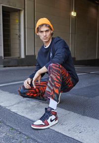 Černá mikina, oranžová čepice, vzorované červené a černé kalhoty, bílé ponožky a černé/červené tenisky Nike; squatuje na chodníku.