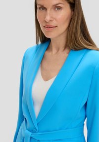 Blazer bleu vif en tissu lisse, avec un col châle, fermeture à un bouton et une ceinture nouée à la taille, superposé sur un haut blanc.