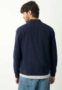 Jaunas vyras su tamsiais garbanotais plaukais, dėvintis tamsiai mėlyną bomber stiliaus striukę ant dryžuotos palaidinės ir mėlynas džinsus, atsisukęs nugarą prieš paprastą foną.