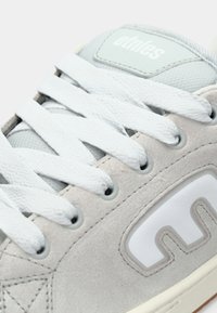 Ανοιχτό γκρι σουέτ αθλητικό παπούτσι με άσπρες κορδόνες, που διαθέτει καουτσούκ σόλα και λογότυπο στη γλώσσα. Παρουσιάζει ενισχυμένες ραφές και υφασμάτικες λεπτομέρειες.