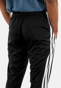 Zwarte sportbroek met witte zijstroken, met een elastische tailleband en een gladde textuur. Toont de achterkant met zakken.