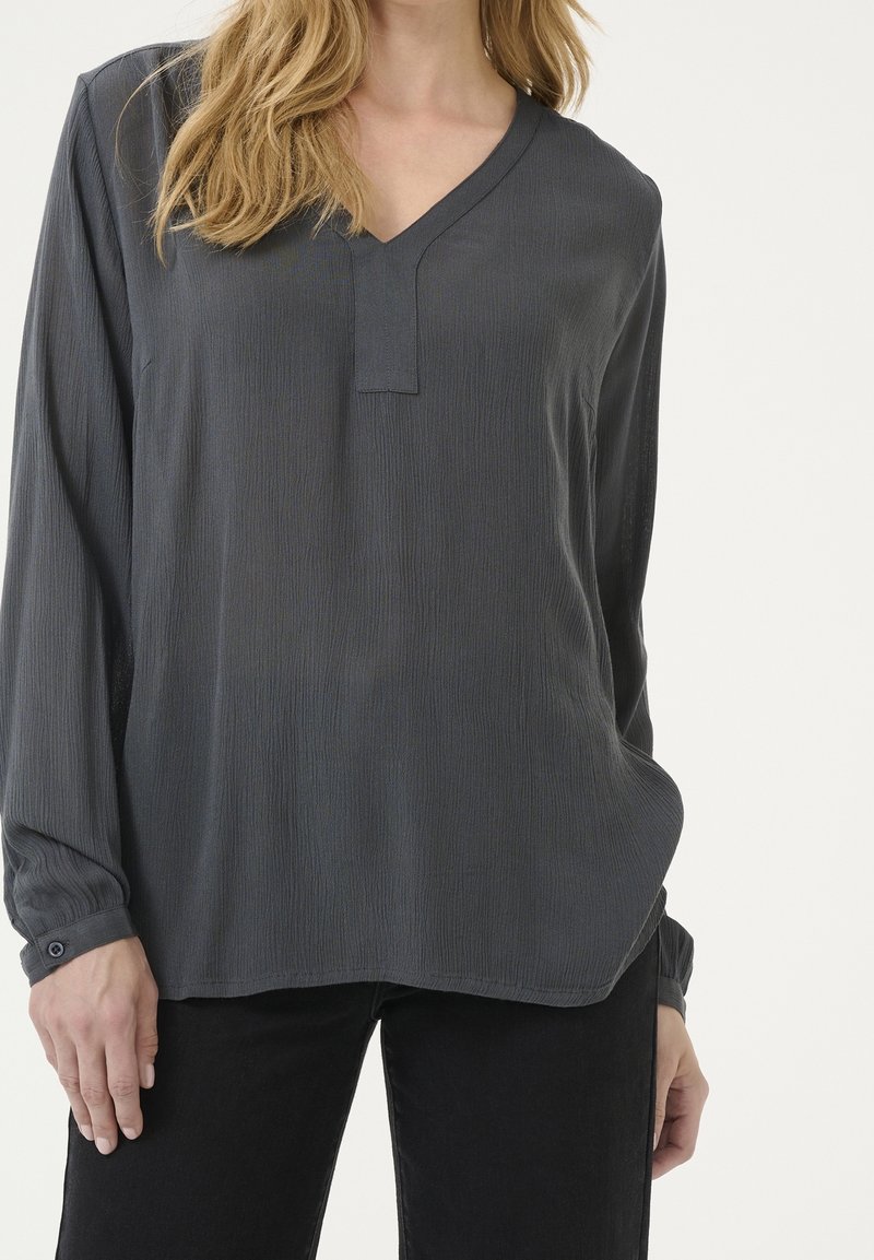 Femme portant un chemisier texturé gris foncé à manches longues avec un col en V et un pantalon noir, debout devant un fond clair uni.