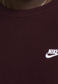 Burgundská pletená svetr s kulatým výstřihem a bílým logem Nike na levé straně. Měkká textura s žebrovaným vzorem.