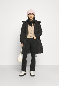 Casaco longo preto acolchoado, suéter de malha bege com botões, calças pretas amplas, botas de tornozelo em creme e um gorro rosa; segurando uma bolsa branca.