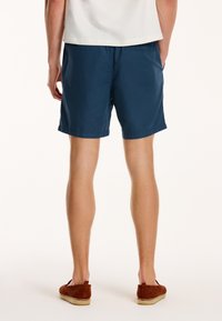 Navy blue cotton shorts with a relaxed fit, featuring an elastic waistband and rolled hem. Worn with brown suede slip-on shoes.