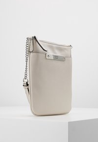 Bolso cruzado beige de Guess con correa de cadena plateada y placa metálica de la marca sobre una superficie blanca.