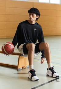Maglietta atletica nera a maniche lunghe con logo dorato, abbinata a pantaloni da basket bianchi e neri. Il modello tiene in mano un pallone da basket rosso e bianco.