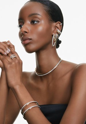 Donna con capelli lisci che indossa molti orecchini a cerchio in oro e argento, un collarino, due braccialetti a polsino e un anello a spirale, su sfondo bianco.