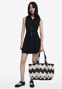Black sleeveless dress with a zip front, belted waist, and side pockets; carrying a large tote bag with black and cream zigzag pattern.