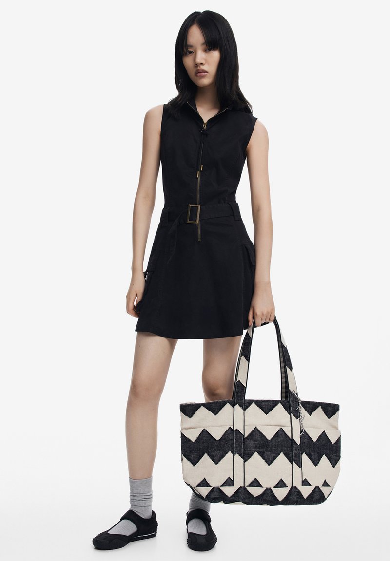 Black sleeveless dress with a zip front, belted waist, and side pockets; carrying a large tote bag with black and cream zigzag pattern.