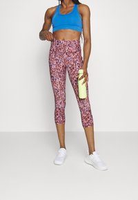 Sportbehå i blått, mönstrade kaprisleggings i rosa och vita sneakers. Leggingsen har ett mjukt, texturerat tyg. Håller en ljusgrön vattenflaska.