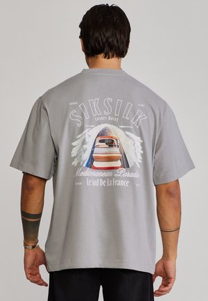 Férfi szürke pólóban, amelyen egy hajó száguld a vízen, alatta a „SIKSILK Luxury Hotel Mediterranean Paradise Le Sud De La France” felirat.
