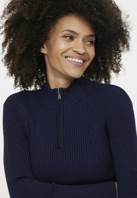 Marineblauer, gerippter Halb-Reißverschluss-Pullover mit hohem Kragen, der über ein strukturiertes Strickmuster und einen silbernen Reißverschluss verfügt. Glattes Material mit einer taillierten Silhouette.