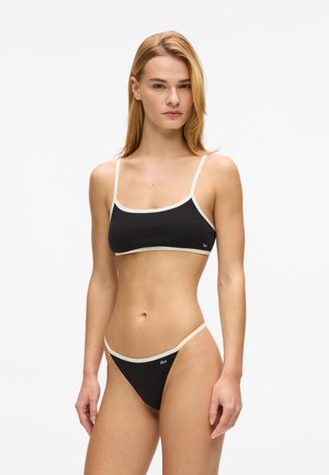 Nuori nainen seisoo musta-valkoreunuksisessa bikineissä, kasvoillaan eteenpäin katsova ilme, valkoisen taustan edessä.