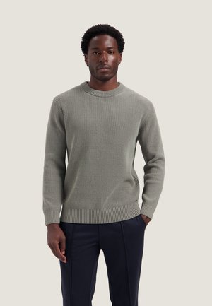 Homme aux cheveux courts et bouclés portant un pull côtelé gris clair et un pantalon foncé, debout avec une main dans la poche.