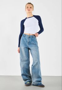 Hvit cropped topp med marineblå langermede, kombinert med løstsittende lyseblå jeans. Modellen har på seg mørke sko med grønne detaljer.