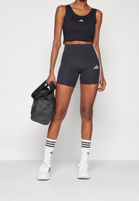 Czarny sportowy crop top i szorty o wysokim stanie z białymi paskami po bokach. Uzupełnione prążkowanymi skarpetkami do kostek i białymi trampkami. Czarna torba w dłoni.