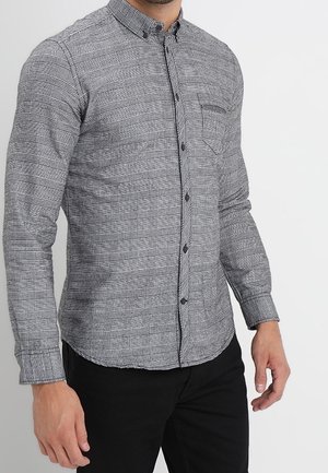 Homme portant une chemise cintrée à carreaux grise à manches longues boutonnée, avec un pantalon noir, debout devant un fond blanc uni.