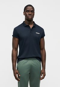 Ναυτικό μπλε μπλουζάκι πόλο με κοντά μανίκια, γιακά και μικρό λογότυπο στο στήθος, συνδυασμένο με ανοιχτό πράσινο παντελόνι με ελαστική μέση.