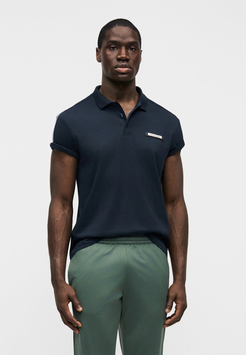 Ναυτικό μπλε μπλουζάκι πόλο με κοντά μανίκια, γιακά και μικρό λογότυπο στο στήθος, συνδυασμένο με ανοιχτό πράσινο παντελόνι με ελαστική μέση.