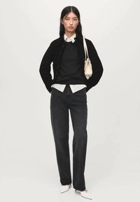 Svart cardigan över svart tröja och vit skjorta, parat med mörka jeans och vita spetsiga skor. Liten handväska i krämfärg som accent.