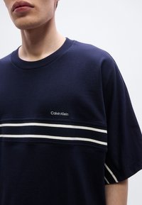 Μπλε ναυτικό βαμβακερό t-shirt με κοντά μανίκια, με λευκή ρίγα στο στήθος και το λογότυπο «Calvin Klein» σε λευκό χρώμα.